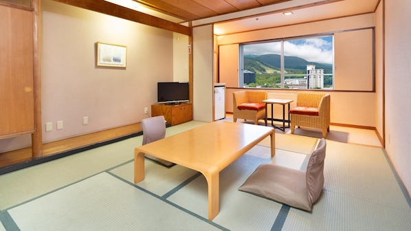 Una stanza tradizionale giapponese con pavimento in tatami, un tavolino, due sedie imbottite, un televisore e una finestra con vista sulle montagne.