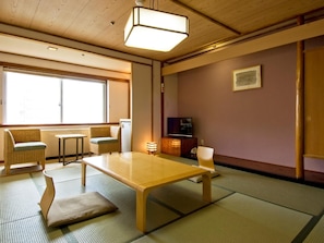Una tradizionale stanza giapponese con pavimento in tatami, un tavolino basso, due poltrone imbottite, un televisore e una porta scorrevole.