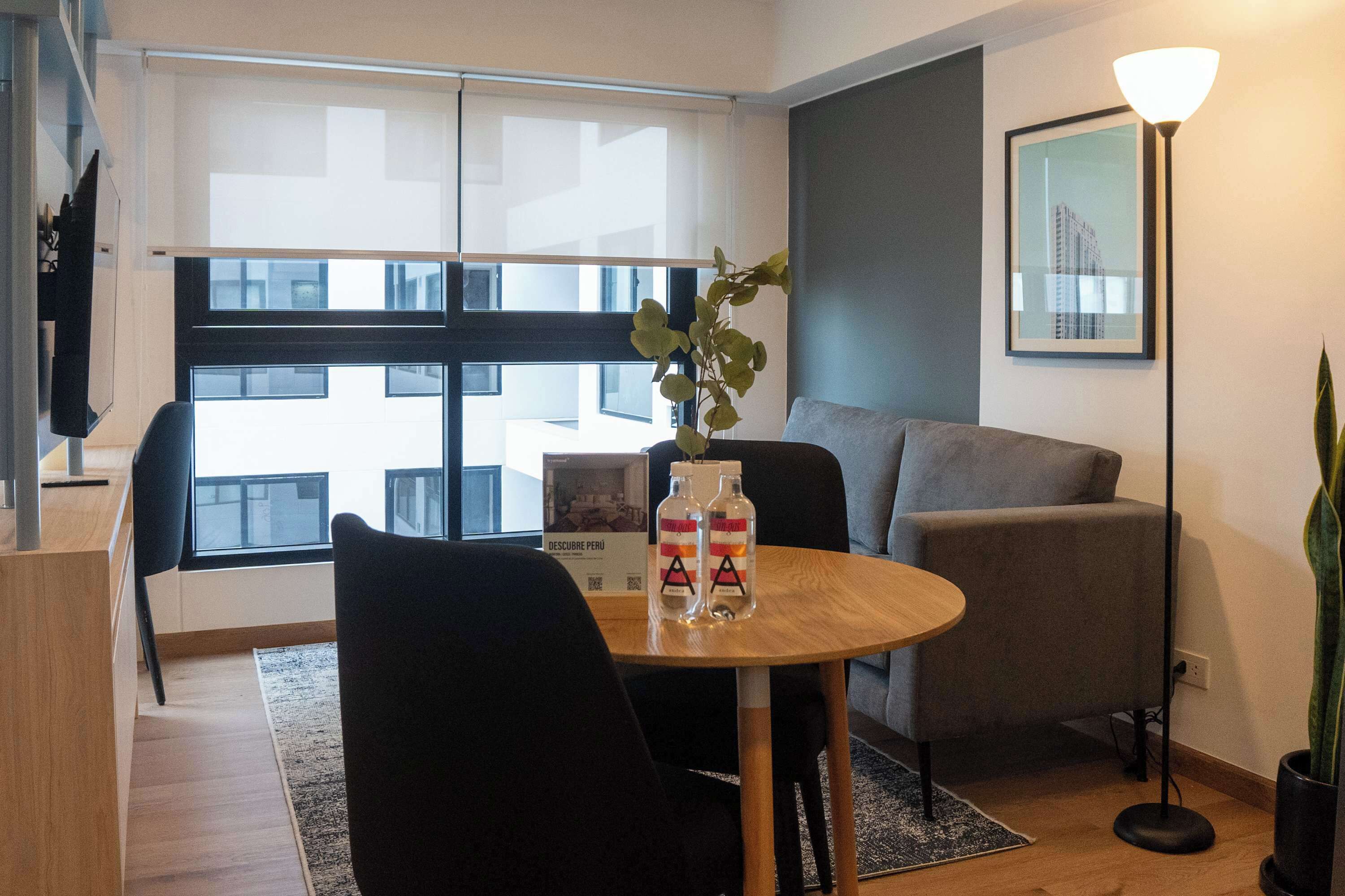 A modern living room with a wooden table, two chairs, a grey sofa, a floor lamp, and a potted plant.