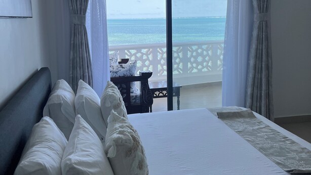 A hotel room with a bed, white pillows, a view of the sea through a large window, and a balcony with a table and chairs. - 2bedroom beachfront Apartmnt Mombasa,Seaview,Ac,pool,gym,restaurant,beach access (Mombasa)