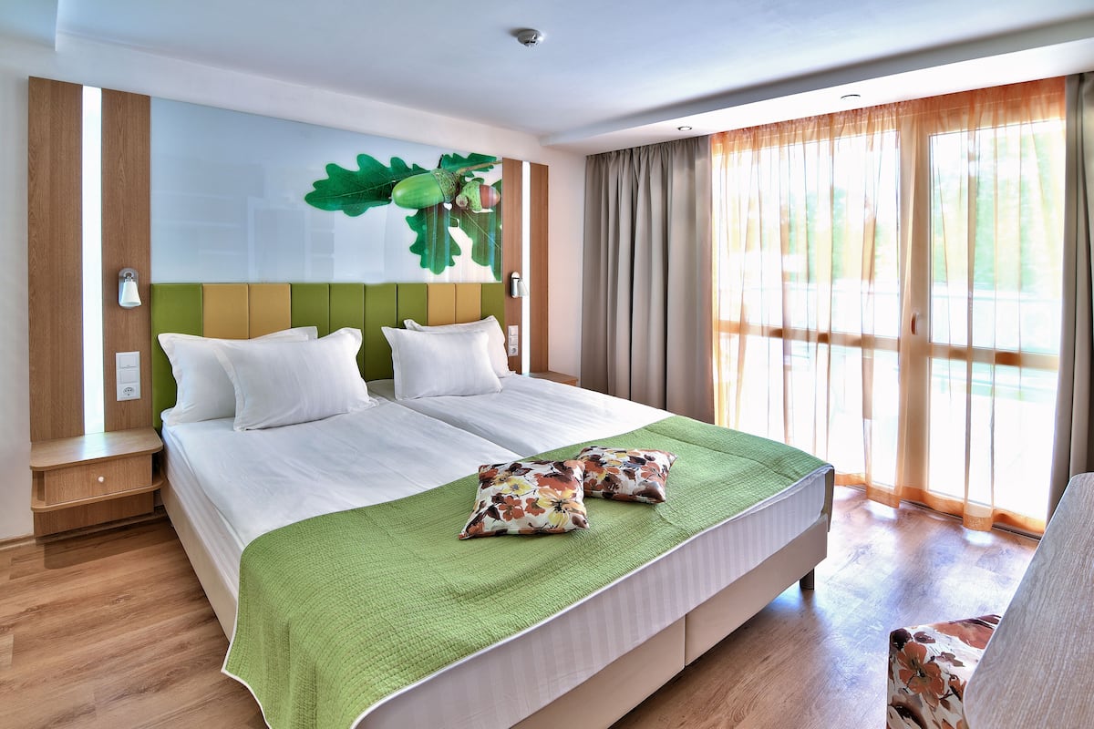 A hotel room with two beds, a green bedspread, and a large window with curtains.