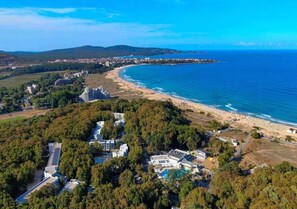 Vista aérea de una zona costera con playa, una zona residencial con edificios modernos y una densa área boscosa.