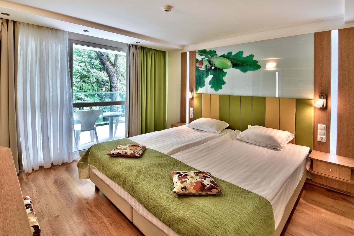 A double bed with green bedding and floral pillows, a nightstand, a wall-mounted lamp, and a large window with curtains.