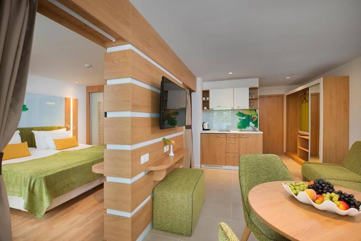A modern hotel room with a bed, a green sofa, a dining table with fruit, and a kitchen area.