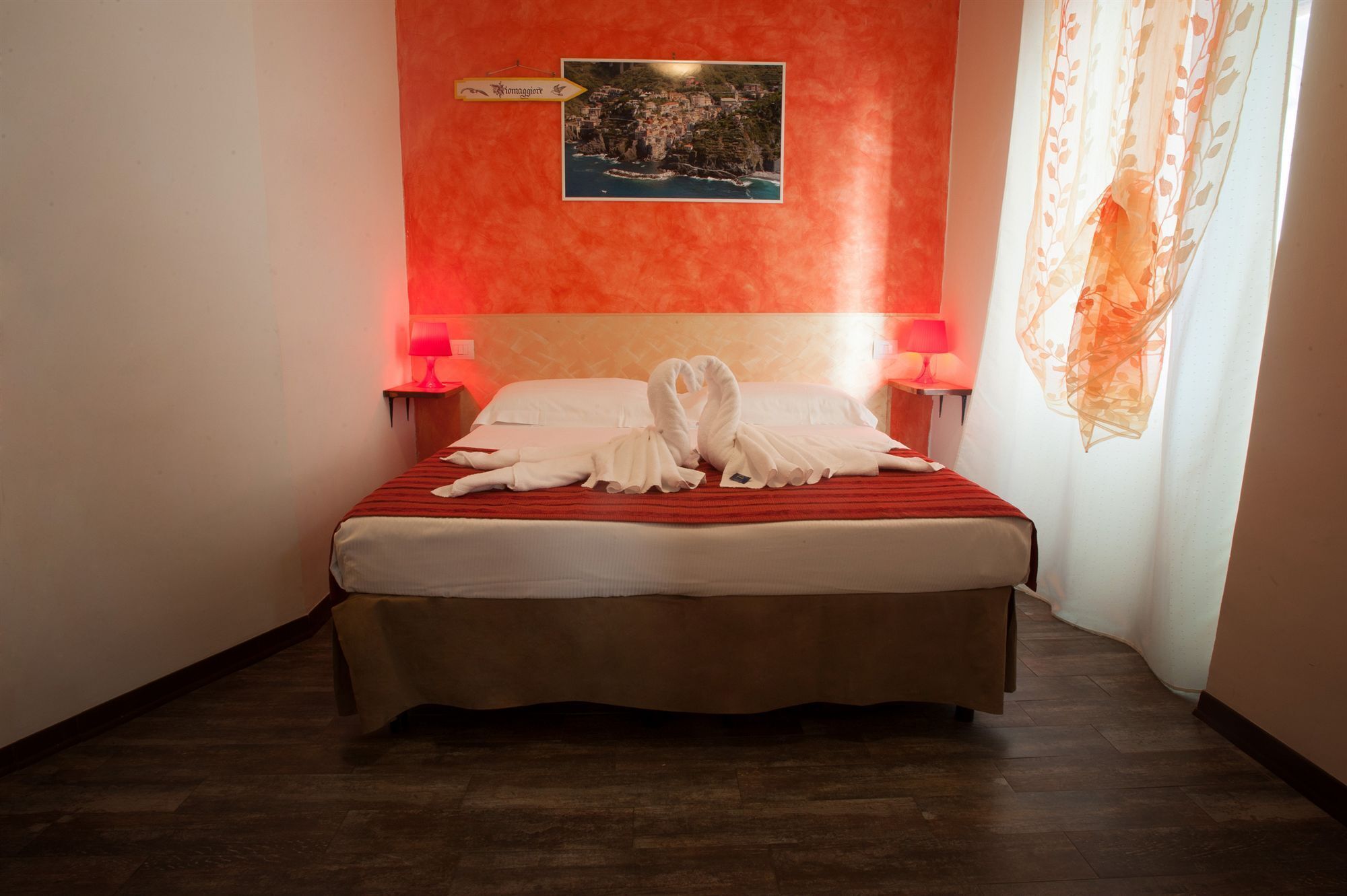 A hotel room with a red wall, a framed picture of a coastal town, two bedside lamps, and a bed with white linens and a red blanket.