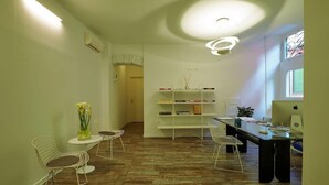 Ein moderner Arbeitsraum mit einem langen Tisch, zwei weißen Stuhlgestellen, einem Bücherregal und einem Computermonitor.
