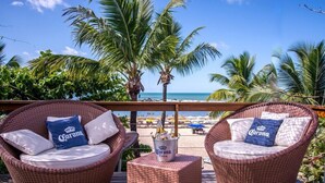 Twee rieten lounge stoelen met kussens en een Corona bieremmer op een houten terras met uitzicht op een strand met palmbomen en parasols.