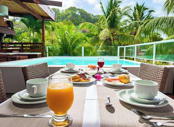 A dining table with breakfast items and a glass of orange juice, overlooking a pool with palm trees and a tropical view. - Sambass Lounge Beach Cafe and Pousada (Cairu)