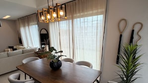 Ein moderner Essbereich mit einem Holztisch, sechs Stühlen, einer Topfpflanze und einem großen Fenster mit Vorhängen.