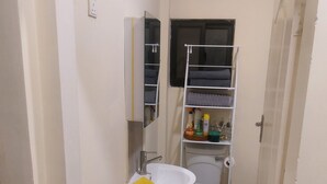 A narrow bathroom with a white sink, mirror, and shelves above the toilet. - 24blouieville (Lusaka)