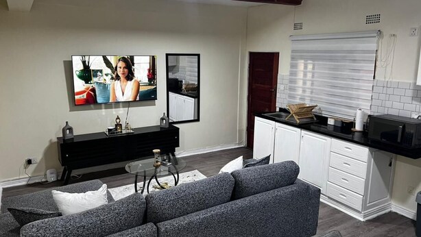 A modern living room with a grey sofa, a black entertainment unit, and a kitchen area with white cabinets and a black countertop. - 24blouieville (Lusaka)