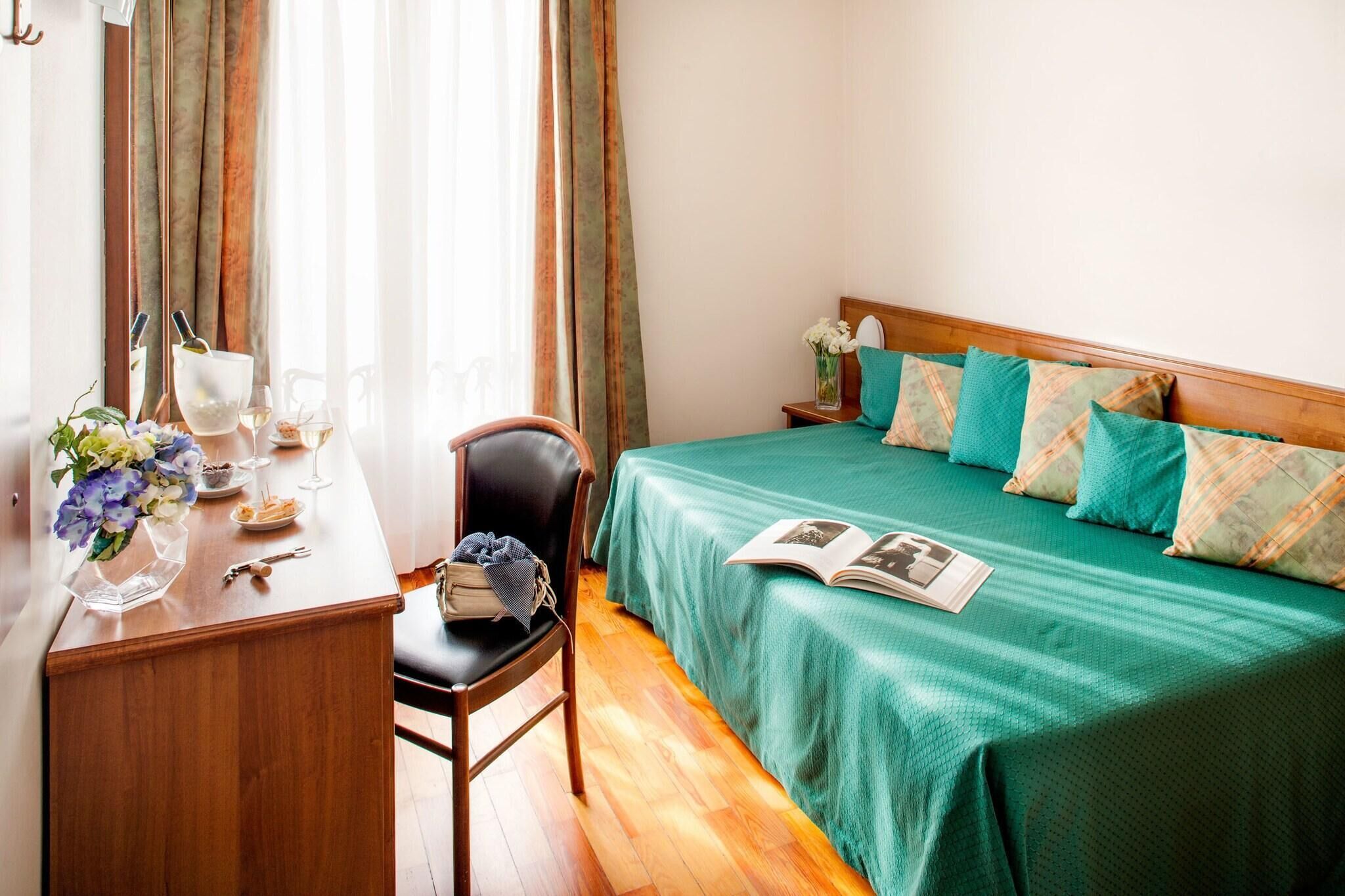 Habitación de hotel con un escritorio de madera, una silla, una cama con ropa de cama verde, una mesita de noche con un jarrón de flores y una ventana con cortinas.
