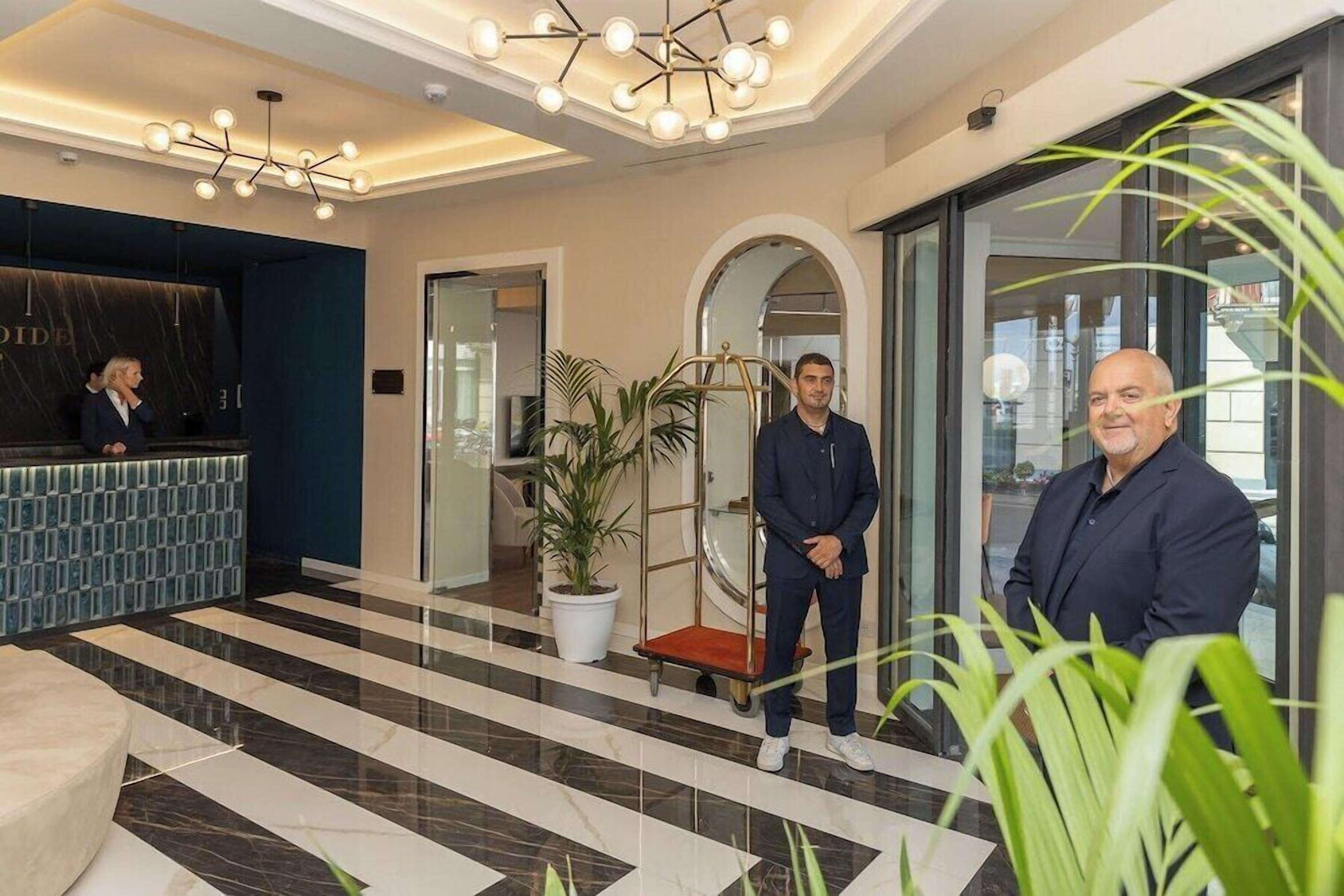 A modern hotel lobby with a reception desk, a large potted plant, and a glass elevator.