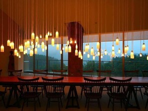 A dining area with a long wooden table and chairs, hanging pendant lights, and a view of the outdoors through large windows. - Furano Hotel (Furano)