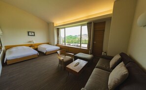 A hotel room with two beds, a sofa, a small table, and a window with a view of greenery. - Furano Hotel (Furano)