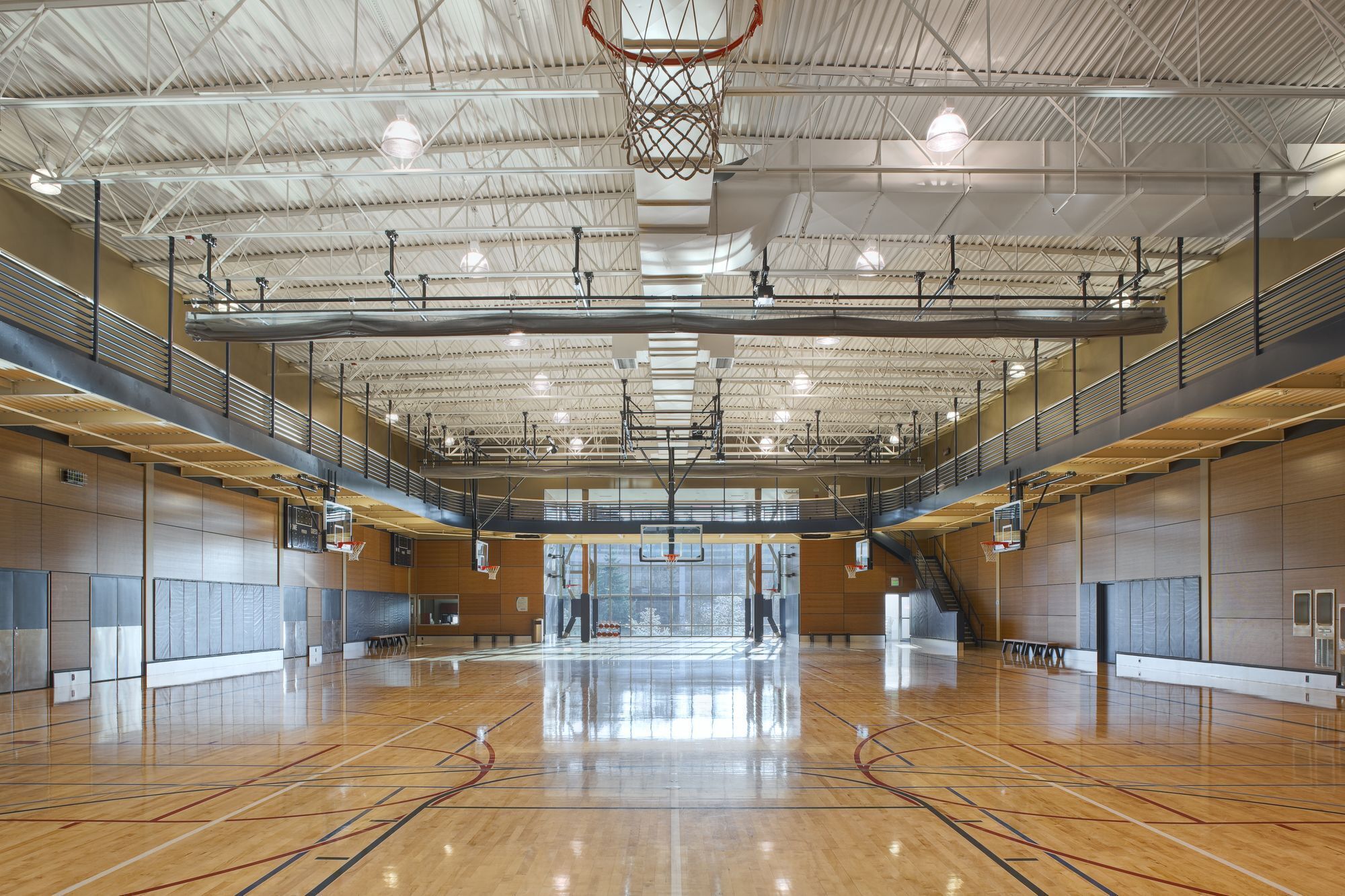 A spacious indoor basketball court with a high ceiling, multiple basketball hoops, and a polished wooden floor.