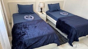 Habitación de hotel con dos camas, cada una con una colcha azul oscuro y almohadas a juego. Entre las camas hay una mesita de noche blanca con una lámpara y una toalla pequeña. La habitación tiene paredes blancas y suelo de mármol.