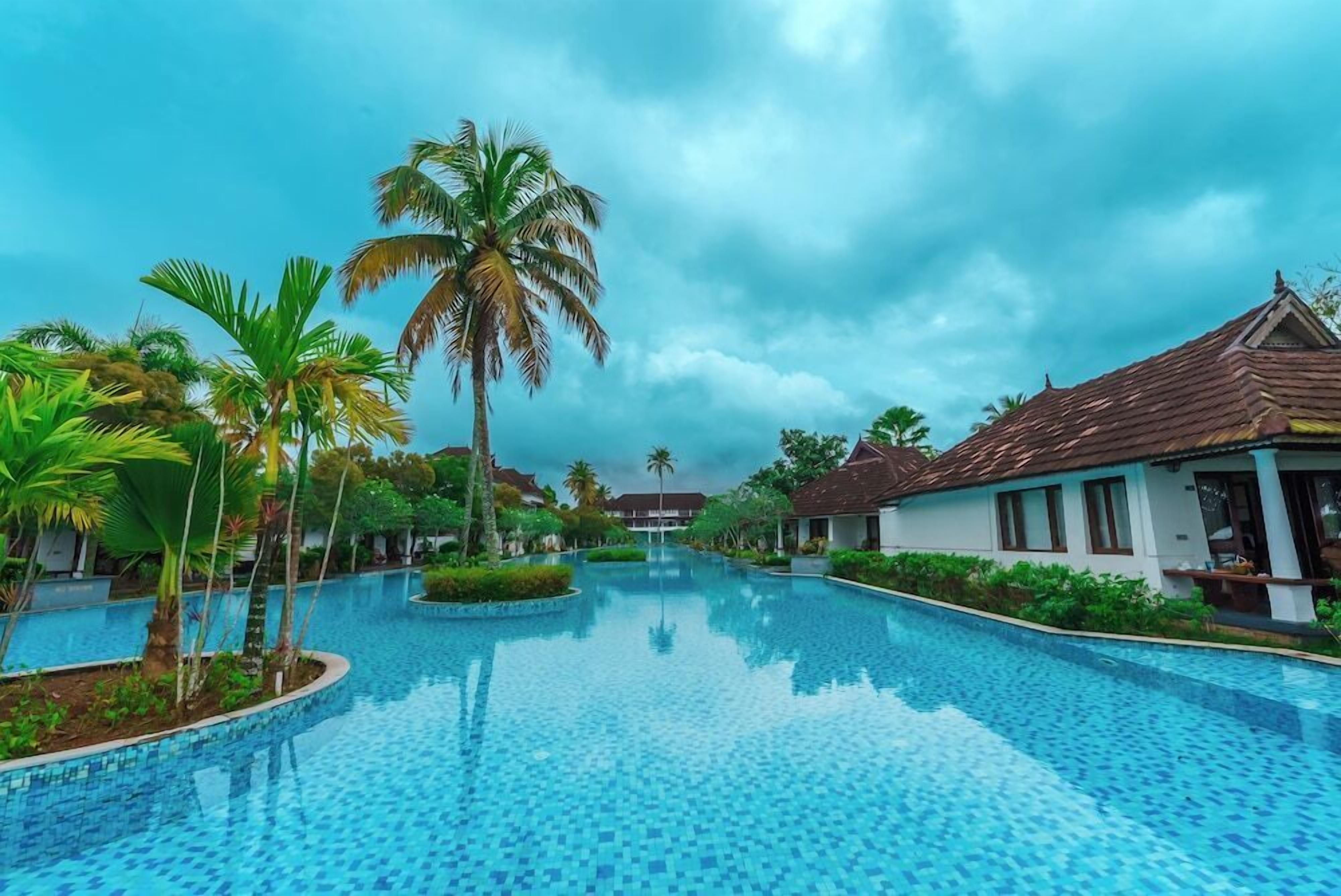 A swimming pool with tiled flooring, surrounded by palm trees and buildings with tiled roofs.