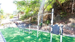 Une salle de sport en plein air, dotée d’appareils d’exercice installés sur une pelouse artificielle, entourée d’arbres et bordée d’une colline rocheuse.