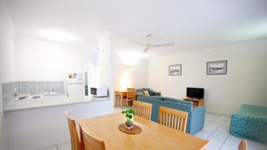 Une cuisine moderne avec des meubles blancs, une table à manger avec des chaises, un four à micro-ondes et un réfrigérateur. Le salon est équipé d’un canapé et d’une télévision.