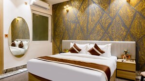 A modern hotel room with a large bed, a ceiling fan, a wall-mounted air conditioner, a bedside table with a lamp, and a decorative mirror. - Hotel Silver Star (Greater Noida)