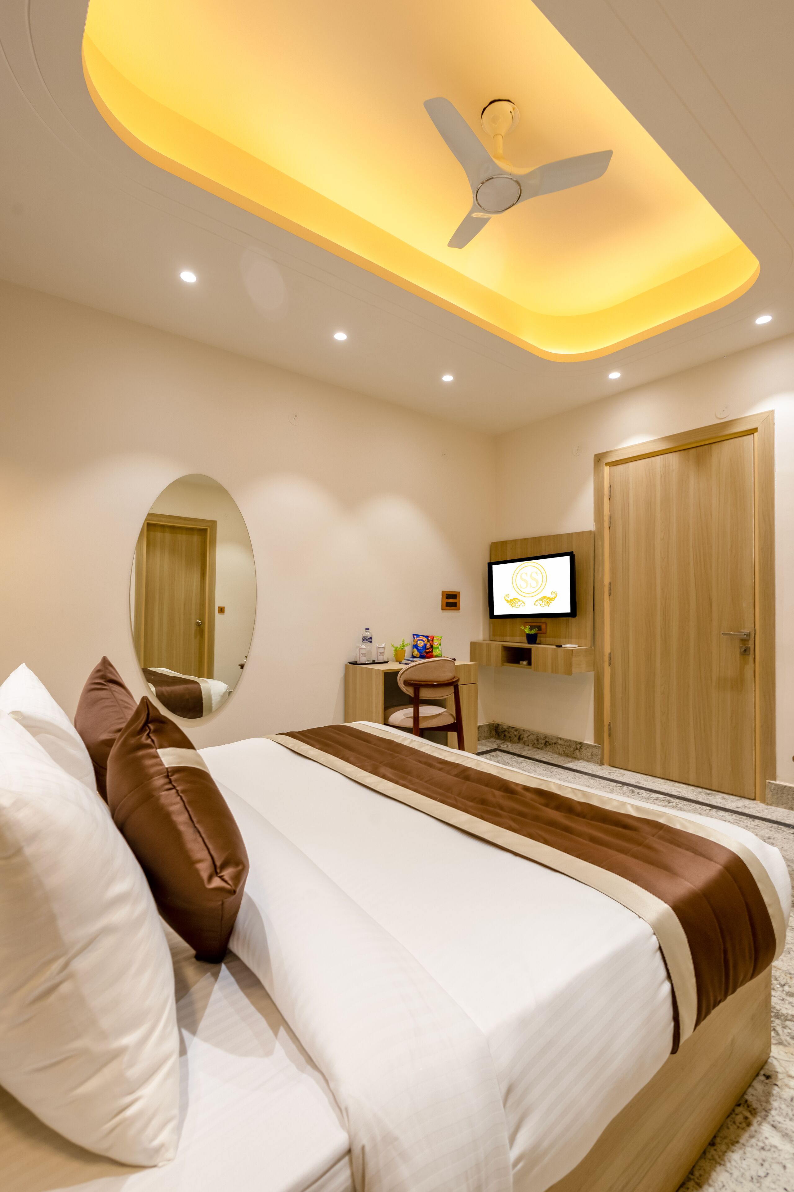 A hotel room with a bed, a ceiling fan, a TV, a mirror, and a wooden door.