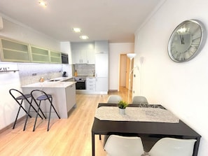 A modern kitchen with a dining area, featuring a large wall clock, a small island with bar stools, and a dining table with chairs. - Renovated beachfront apartment in Roses, air conditioning and elevator (Roses)