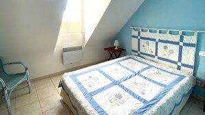Une chambre avec un plafond en pente, un lit recouvert d’une couette bleue et blanche, une chaise, une petite table avec une lampe et une fenêtre avec un radiateur.