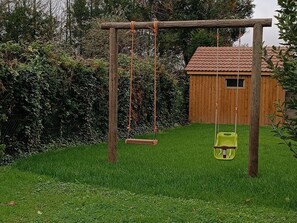 Een houten schommel in een gazon met een houten tuinhuis op de achtergrond.