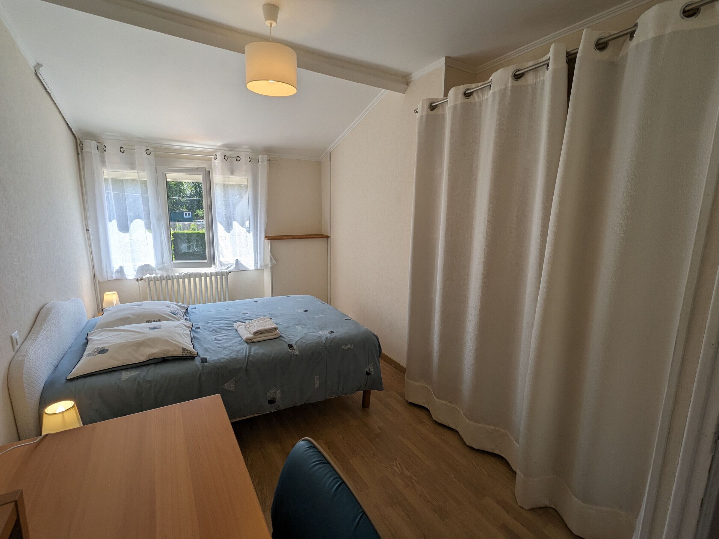 A bedroom with a bed, a desk, a chair, a window with curtains, and a radiator.