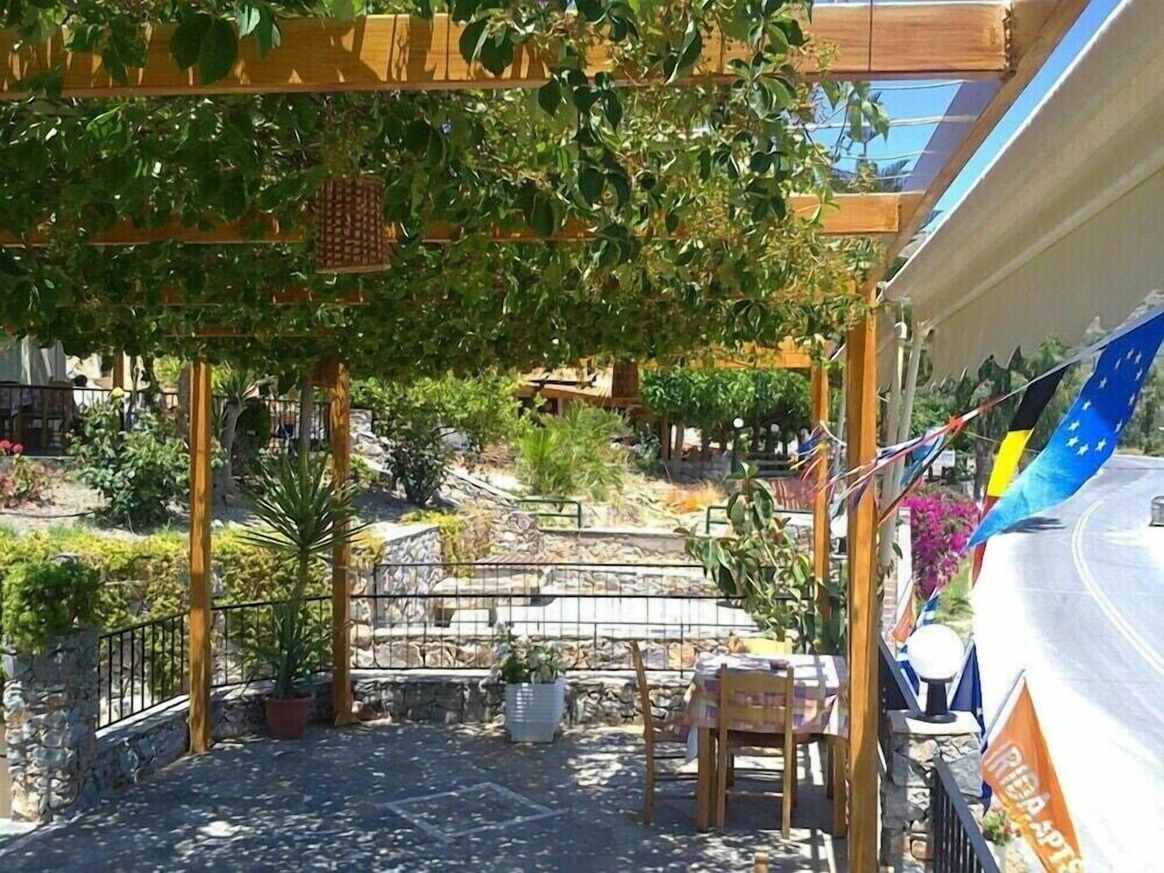Un espacio al aire libre cubierto con una pérgola, plantas en macetas, una mesa con sillas y banderas.