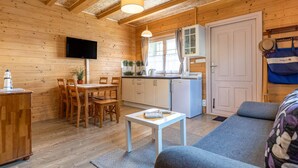 Una habitació acollidora d'estil cabana amb interior de fusta, zona de dinar amb taula i cadires, petita cuina amb nevera i rentatges, i una finestra gran que proporciona llum natural.
