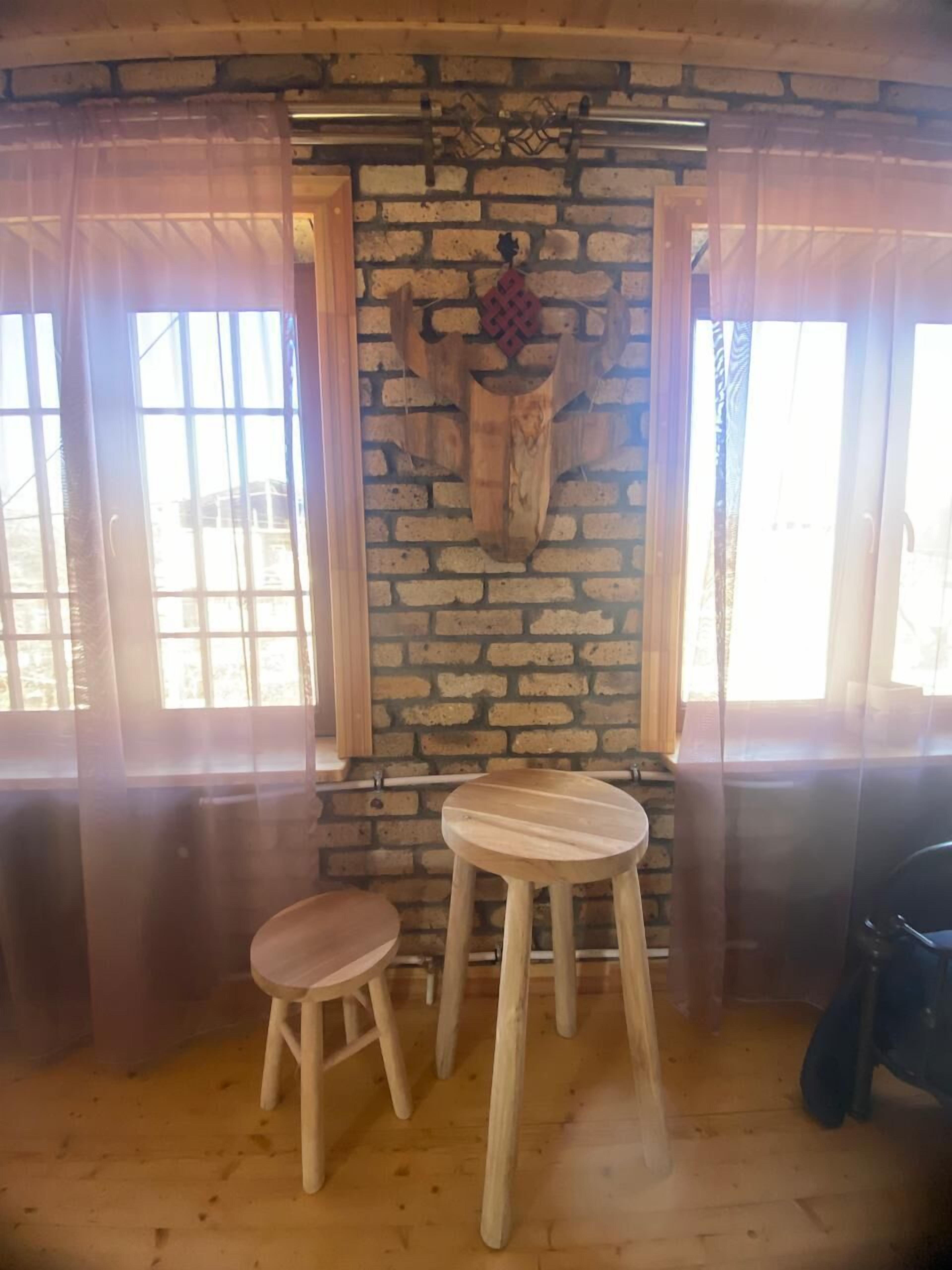 Een rustieke kamer met een bakstenen muur, een houten tafel en kruk, en een groot raam met gordijnen.