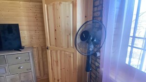 A room with wooden walls and ceiling, a wooden cabinet with drawers, a standing fan, and a window with curtains.