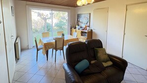 Un salon avec un plafond en bois, un coin repas avec une table et des chaises, un canapé avec des coussins et une vue dégagée sur l’extérieur grâce à une grande fenêtre.