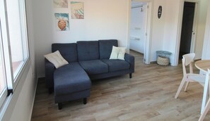 A modern living room with a dark grey sectional sofa, two white pillows, a small round table, and a white chair. There are wooden floors and a door leading to another room. - Modern Apartment in Palamós, with Air Conditioning (Palamós)