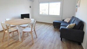 Ein Wohnzimmer mit einem runden Holztisch, vier weißen Stühlen, einem dunkelgrauen Sofa mit zwei Kissen und einem Fenster mit Blick auf Gebäude.