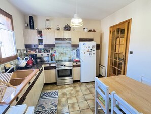 A compact kitchen with a stove, oven, refrigerator, and a dining area with a table and chairs. - Vacation Home Near the Lake (Biscarrosse)