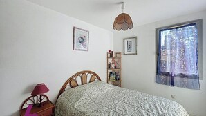Een slaapkamer met een bed, een nachtkastje met een lamp, een boekenkast en een raam met uitzicht op bomen.