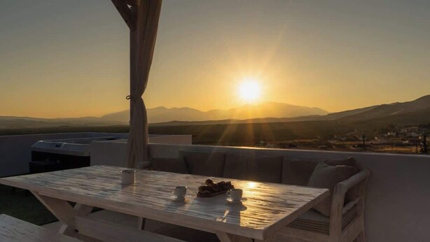Una terraza con mesa y sillas, una parrilla y vistas a la puesta de sol sobre montañas.