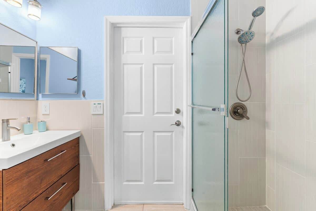 Un baño con puerta blanca, cabina de ducha con mampara de vidrio y tocador con espejo y lavamanos.
