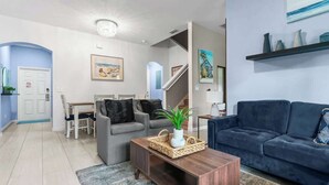 Ein modernes Wohnzimmer mit einem blauen Sofa, einem grauen Sofa, einem Holztisch und einem Essbereich mit weißem Tisch und Stühlen.