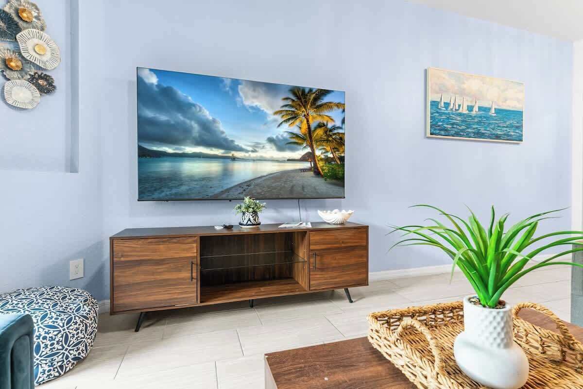 Een woonkamer met een grote flatscreen televisie aan de muur, een houten kast, een ottoman met een patroon en een kamerplant.