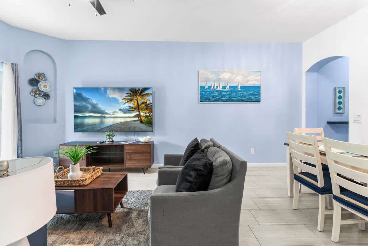 A modern living room with a grey sofa, a coffee table, and a TV. There are paintings on the wall and a dining area with wooden chairs.