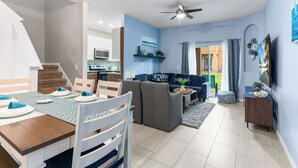 Ein modernes Wohnzimmer mit einer Couch, einem Esstisch und einem Deckenventilator, im Hintergrund eine Küche.