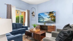 Ein Wohnzimmer mit einem dunkelholztischen Couchtisch, einem blauen Sofa, einem grauen Sessel und einem großen Flachbildfernseher an der Wand.