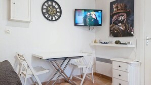 Une petite pièce comprenant un bureau blanc, une télévision, une horloge et un tableau représentant un chat portant un chapeau.