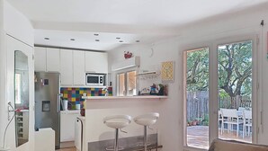 Une cuisine compacte équipée d’un réfrigérateur, d’un four à micro-ondes et d’un plan de travail coloré. On y trouve deux tabourets de bar et un coin repas visible à travers une porte vitrée.