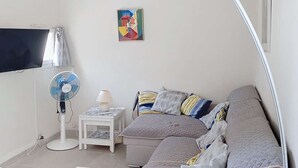 Ett litet, väl upplyst rum med en soffa, ett sidobord med en lampa, en golvfläkt och en tv som är monterad på väggen.
