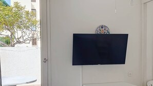 Ett rum med en platt-TV som är monterad på väggen, ett litet vitt skåp under den och ett fönster med utsikt över träd och en byggnad utanför.